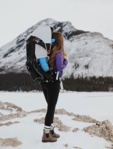 A mom wears an Osprey hiking pack, with her child nestled inside, while hiking in the winter.