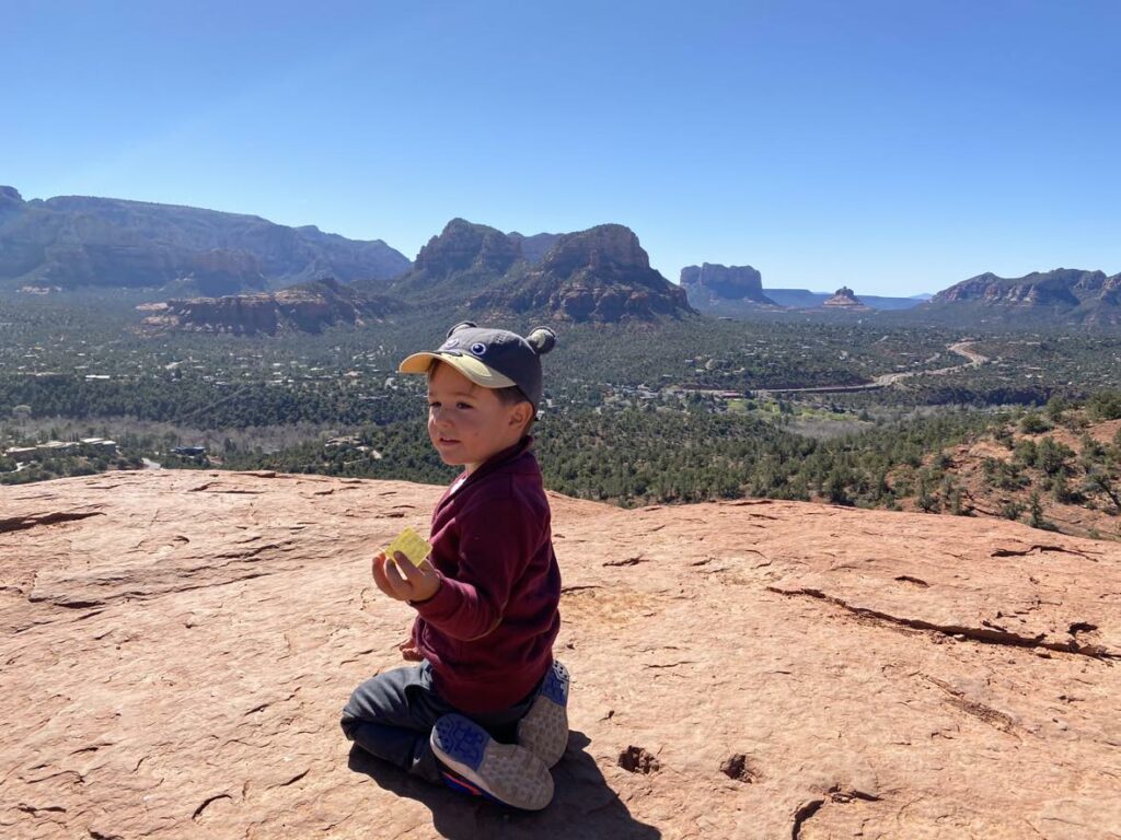A young boy sits along a hiking trail with a sweeping view of Sedona's iconic red rocks in the distance.