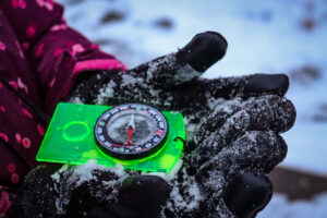 Two gloved hands hold out a green compass, one of the winter hiking essentials needed with kids.