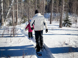 A dad and his young daughter snowshoe through the woods.