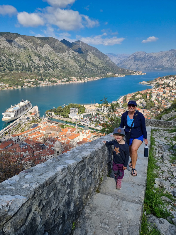 A woman and her young child trek across an old city wall in Montenegro, with the ocean in the distance.