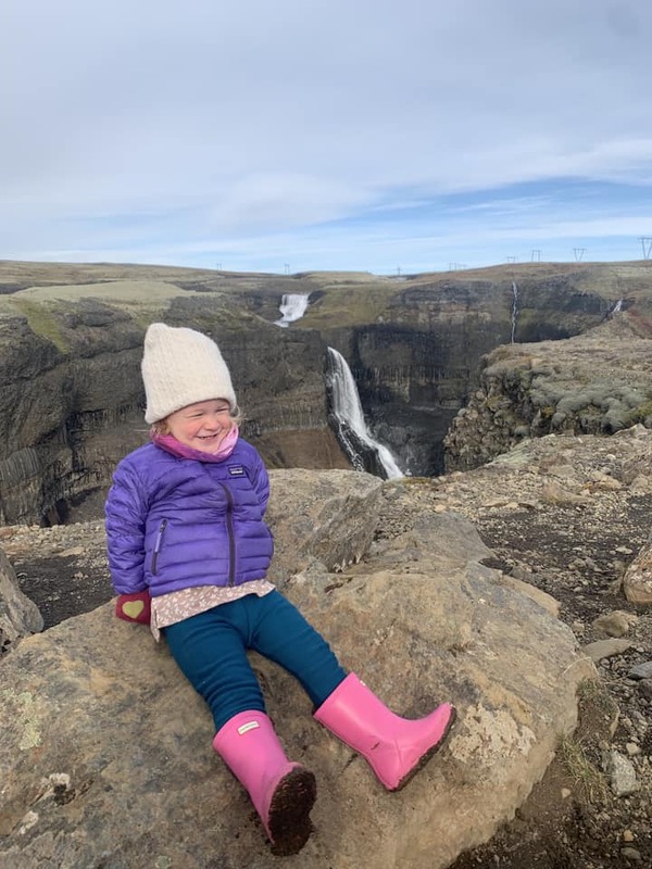 A young girl sits on a rock with a waterfall in Iceland in the distance.