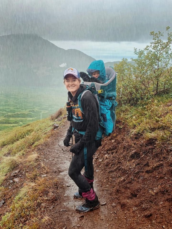 A mom carriers her baby on her back while hiking in the rain. Learning safety tips is a keep part of knowing how to hike in the rain with kids.